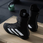 Black Adidas boxing boots on a wooden box with gym equipment in the background