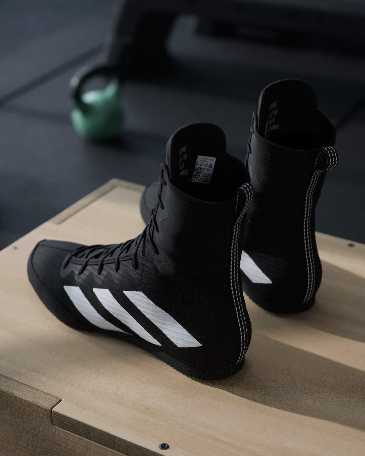 Black Adidas boxing boots on a wooden box with gym equipment in the background